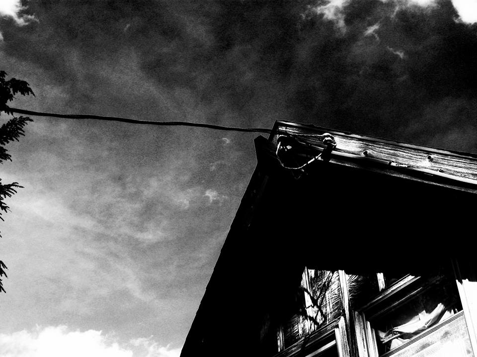 A high-contrast black-and-white image of the tip of a house where the powerline meets. Domineering and looming.