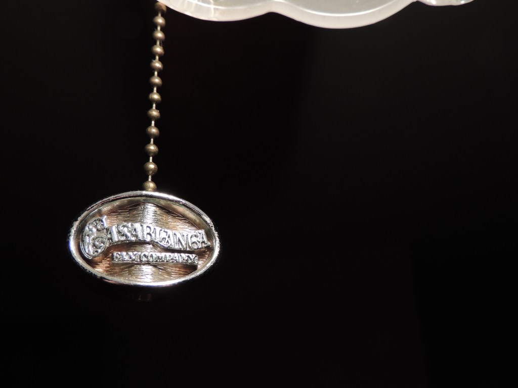 The pull chain on the ceiling fan at my partner's house. A worn metal oval with "CASA BLANCA FAN COMPANY" written across it. In this photo, it's a bit further away, with the white lighting from the fan making the background almost black, highlighting the text on the pendant of the chain.